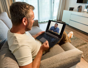 White American person attending a virtual dental consultation from home, sitting on a couch with a laptop in a bright, modern living room.